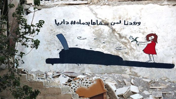A poignant mural by Abu Malik al-Shami on the crumbling wall of a war-torn building in Darayya, Syria. The artwork features a young girl in a red dress standing at a grave, holding a white flower. The tombstone is simple and somber, with petals and leaves scattered around it, symbolizing loss and remembrance. Above the scene, Arabic text translates to "Our flowers, for whom we watered them with our blood #<a href=