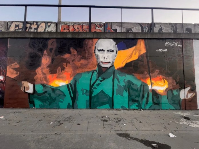 Street art mural by Polish artist Kawu in Wilda, Poznań, depicting Vladimir Putin as Lord Voldemort from the Harry Potter series. The artwork features Putin with pale skin, piercing eyes, and a cloak, surrounded by flames and destruction, symbolizing chaos and fear. The background includes the Ukrainian flag, emphasizing the mural's anti-war message and solidarity with Ukraine. The hashtag #<a href=