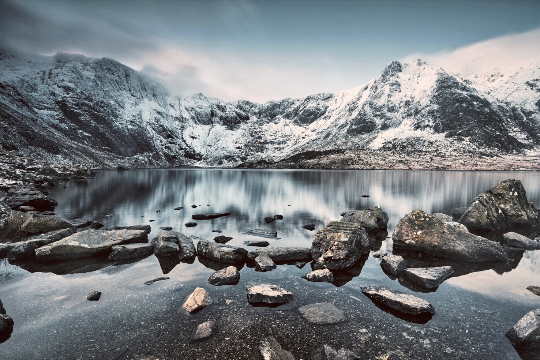 Before sunrise at Cwm Idwal. The mountains are covered with snow. The lake shimmers in the early light. Rocks in the foreground and mountains fill the background beyond the lake.