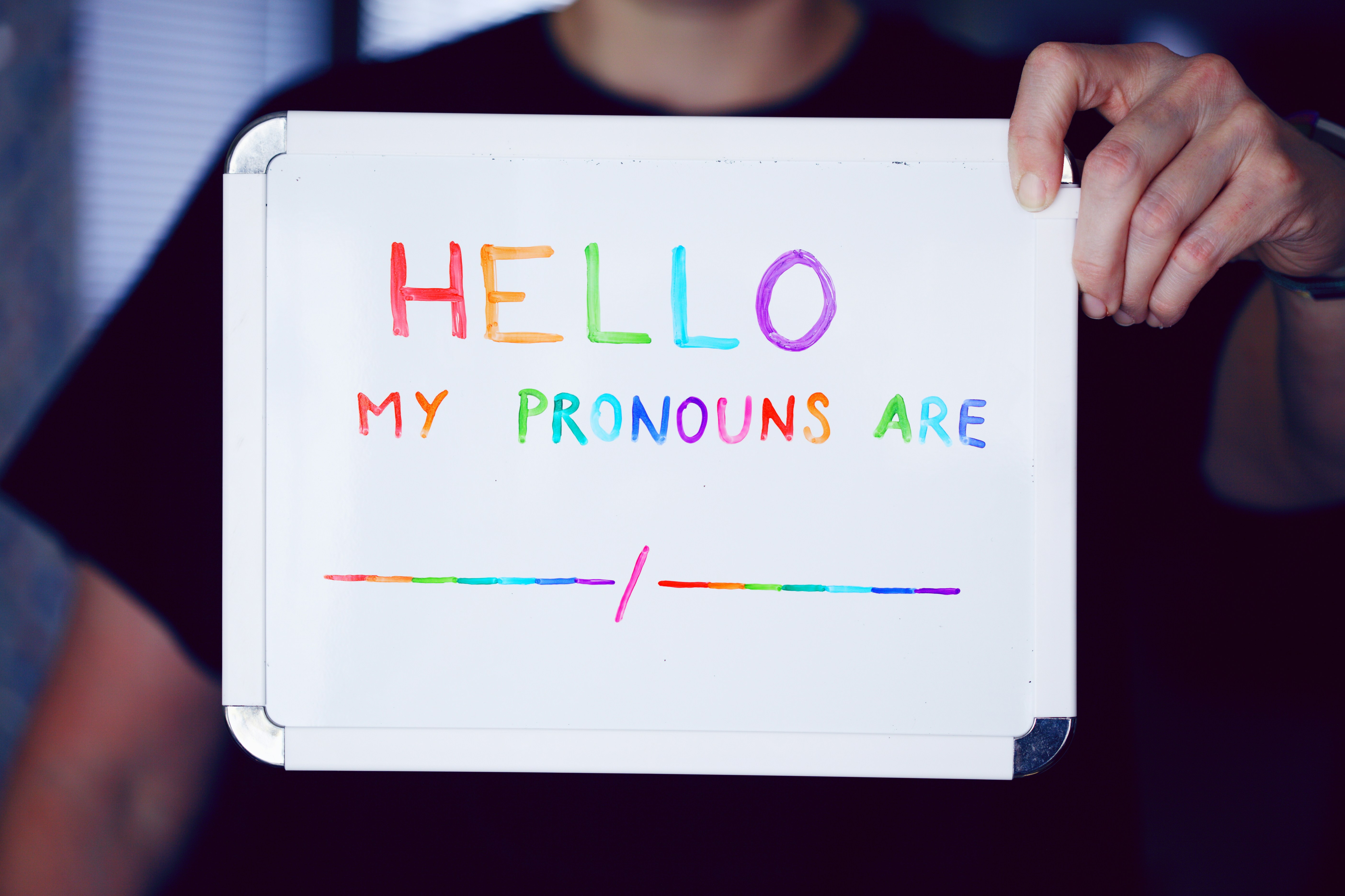 A person holding a whiteboard with their hand. The whiteboard has a message written in multi-colored letters that reads "HELLO MY PRONOUNS ARE" followed by a space with a line underneath for the person to fill in their preferred pronouns. The background is dimly lit, and the focus is on the whiteboard and the person's hands.