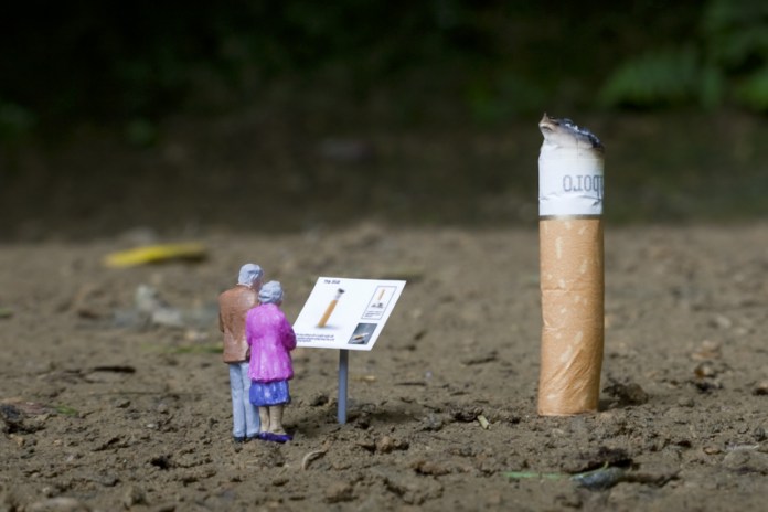 Artwork from the 'Little People' series by Slinkachu, featuring two miniature figurines, an elderly man and woman, standing in front of a sign resembling a museum exhibit description. The sign explains a cigarette butt, which looms large in comparison to the tiny figures, turning the discarded object into an oversized, thought-provoking display. The scene is set on a patch of dirt, blending urban commentary with whimsical scale manipulation.