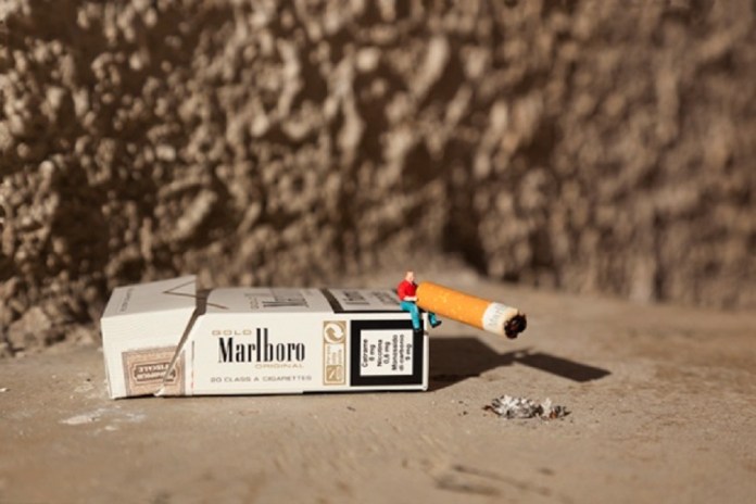 Another piece from Slinkachu's 'Little People' series, showing a miniature figurine sitting on a cigarette butt, which rests on an opened Marlboro cigarette pack. The small figure, dressed in a red sweater and blue pants, interacts with the oversized objects, highlighting themes of consumption and waste. The scene is set against a textured wall, with a small pile of ash nearby.