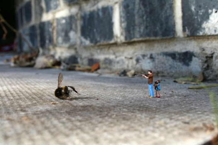 A whimsical scene from Slinkachu's 'Little People' series, featuring two miniature figurines—one holding a rifle, the other observing—standing on a sidewalk in front of a dead bumblebee. The oversized bee transforms into a dramatic, almost otherworldly creature in this context, while the gray stone wall in the background adds a gritty, urban atmosphere.