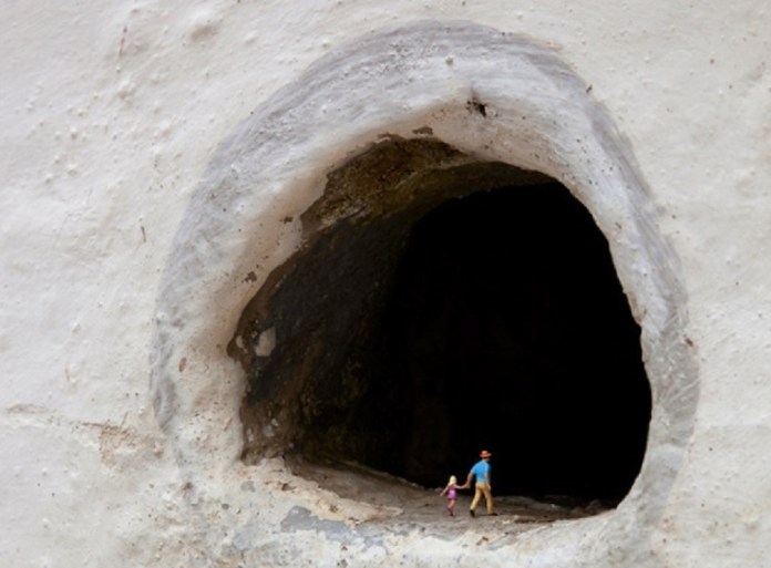 A piece from Slinkachu's 'Little People' series, featuring two miniature figurines—a man in a blue shirt and hat holding hands with a small child in pink—standing at the edge of a large, dark hole in a textured white wall. The contrast between the figurines' bright colors and the ominous darkness of the hole creates a sense of adventure and mystery.