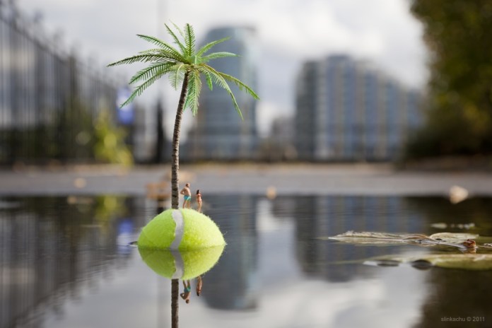 A whimsical installation from Slinkachu's 'Little People' series, featuring two miniature figurines standing on a tennis ball floating in a puddle, styled as a tropical island. A small plastic palm tree rises from the tennis ball, creating the illusion of a deserted island. The background includes blurred urban structures, contrasting with the playful and imaginative foreground.