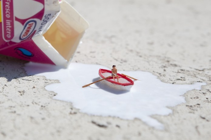 A creative piece from Slinkachu's 'Little People' series, showing a miniature figurine rowing a tiny red boat across a puddle of spilled milk. The milk originates from a toppled 500ml carton lying in the background. The scene transforms a simple spill into a playful and imaginative micro-world.
