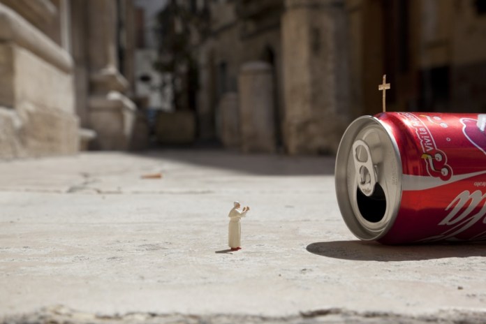 A piece from Slinkachu's 'Little People' series, featuring a miniature figurine of a priest or pope standing in front of a tipped-over soda can with a small cross planted on its top. The scene humorously resembles a religious moment or ceremony, with the soda can acting as an improvised altar. The background shows a blurred urban alley, contrasting with the playful foreground.