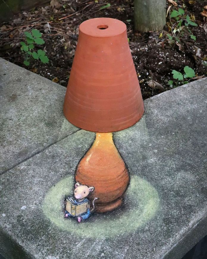 A delightful chalk art piece by David Zinn titled "Nadine’s Evening of Adventure." The artwork cleverly integrates a real clay flower pot, positioned upside-down, to form the lampshade of a glowing lamp. Beneath the lamp, a tiny chalk-drawn mouse named Nadine sits comfortably, engrossed in reading a book. The scene is painted on a concrete surface, with nearby soil and greenery adding to the cozy ambiance. Zinn's mastery creates a whimsical story, blending real objects and artistic imagination seamlessly.