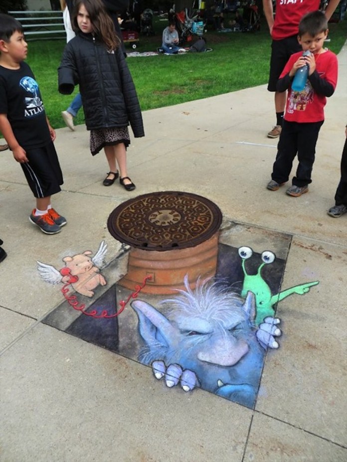A lively chalk art scene by David Zinn depicts a whimsical interaction emerging from a manhole cover on a sidewalk. A blue troll with shaggy hair peeks out, grasping the edge of the hole, while a green alien with antennae stands beside him, appearing amused. A small winged pig holds a red string phone, extending the playful narrative. The art seamlessly integrates a real manhole cover, enhancing the 3D illusion. Children gather around, adding a sense of curiosity and engagement to the scene, set against a backdrop of grass and park visitors.