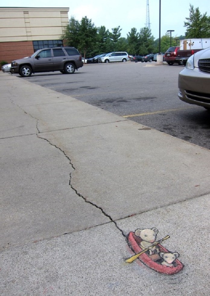 A charming chalk art piece by David Zinn turns a sidewalk crack into a playful scene. The crack is reimagined as a river, where two adorable mice paddle a red canoe with yellow oars. The larger mouse confidently steers while the smaller one enjoys the ride. The setting includes a parking lot with cars, trees, and a building in the background, blending the whimsical artwork seamlessly with the everyday urban environment. Zinn’s creativity brings a touch of delight to the mundane