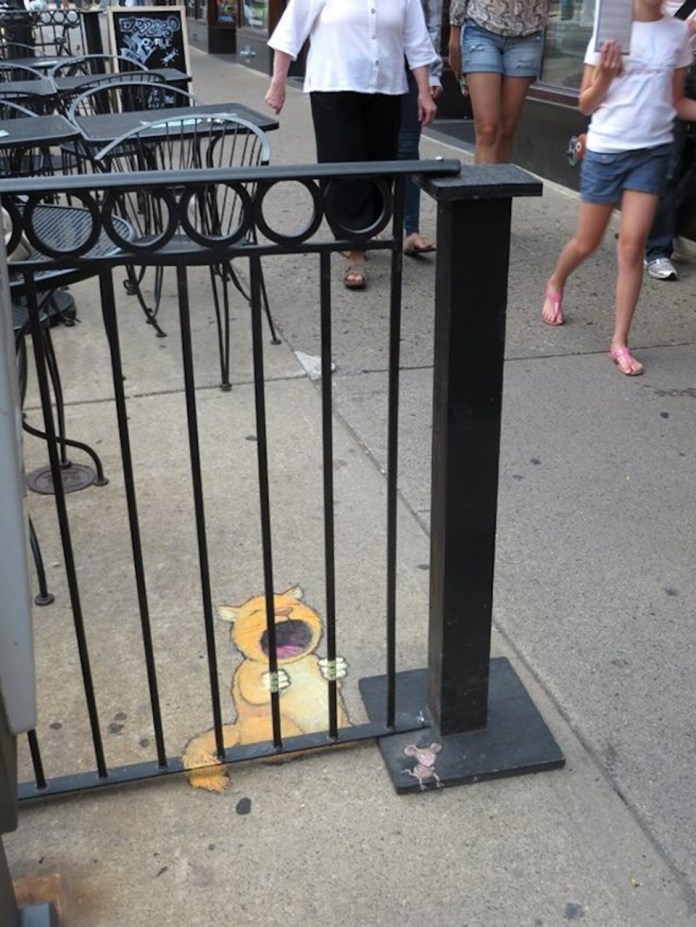 A playful chalk art piece by David Zinn, depicting a cartoonish orange creature seemingly trapped behind the bars of a black metal fence. The creature's exaggerated expression shows it yelling or crying, adding a comedic touch. Nearby, a small chalk-drawn mouse stands on the base of the fence, observing the scene. The artwork is integrated into the urban sidewalk outside a café, with metal tables and chairs visible in the background. Passersby walk past, adding a sense of movement and liveliness to the setting.