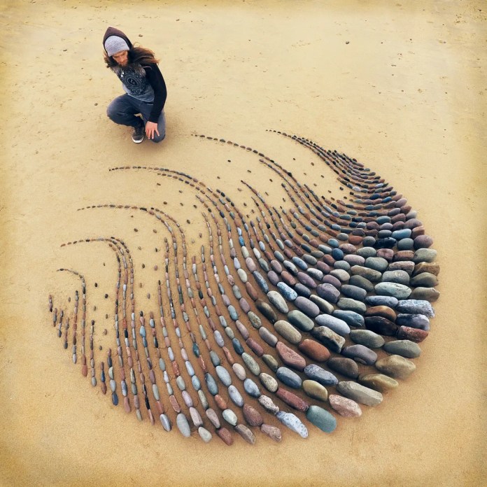 A land art creation by Jon Foreman on a sandy beach. The artwork features a mesmerizing pattern of smooth, colorful stones arranged in swirling waves that taper off into delicate lines of smaller stones. Foreman is kneeling nearby, wearing a hoodie and beanie, adding a human connection to the ephemeral artwork.