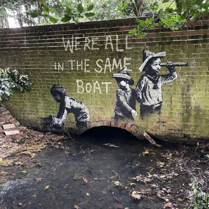 A Banksy mural titled "We're All in the Same Boat" on a mossy brick wall above a small waterway in Oulton Broad, England. The artwork features three children in monochrome: one crouching to reach into the water, one standing and holding a telescope, and one looking ahead, all wearing paper hats. The text "WE'RE ALL IN THE SAME BOAT" is painted in white above them, blending with the natural greenery around the wall.
