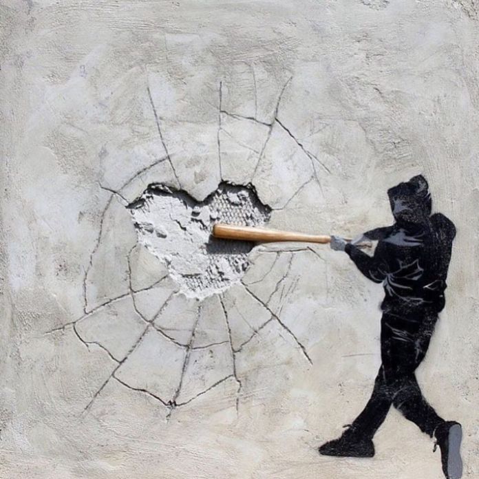 Stencil-style street art by Hijack showing a figure in a black hoodie swinging a baseball bat into a concrete wall. The bat appears to have cracked the surface, revealing a heart-shaped hole with fractured lines radiating outward