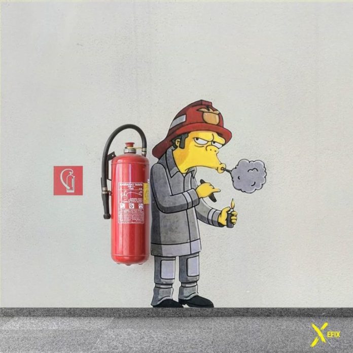 A street art piece by EFIX featuring a firefighter character from The Simpsons, humorously interacting with a real fire extinguisher mounted on the wall. The painted firefighter holds a lit cigarette, exhaling smoke, creating an ironic and playful contrast with the safety equipment. The artwork cleverly incorporates the fire extinguisher into the scene, set against a plain white wall with a fire safety sign nearby.