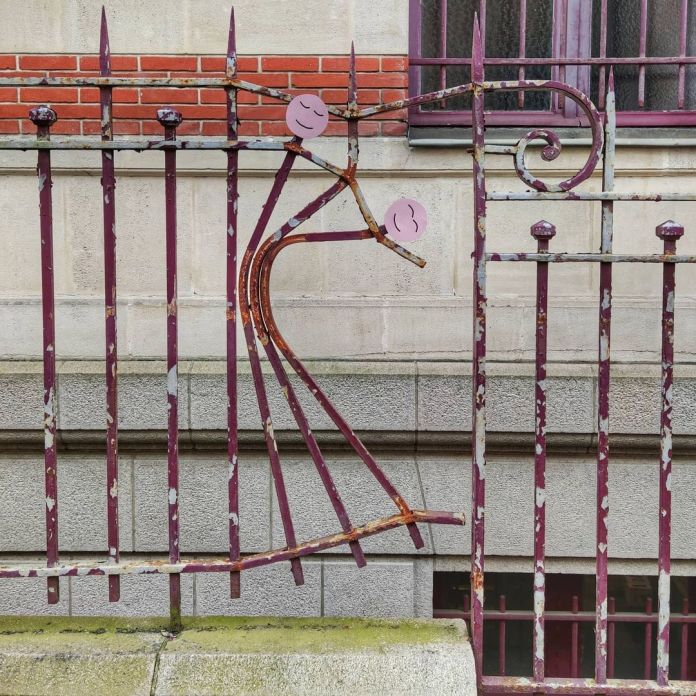 A charming street art piece by Oakoak that transforms a rusted section of an old iron fence into a playful depiction of a figure. The bent metal bars form the flowing body of a dancing person, while two painted pink circles with smiling faces serve as the head and hand. The artwork is set against a background of a brick and stone building, blending the urban decay of the fence with a whimsical narrative. This piece exemplifies Oakoak's knack for turning overlooked urban details into delightful and imaginative artworks.