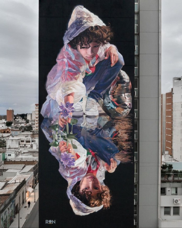 A towering mural by Martín Ron titled "Reflections," located in San Nicolás de los Arroyos, Argentina. The artwork depicts a young person crouching, wearing a translucent plastic raincoat with vibrant reflections, holding purple flowers. The figure’s mirrored image appears below, seamlessly blending into the surface. This hyper-realistic piece, rich in detail and color, occupies the side of a tall urban building, contrasting against the muted cityscape under a cloudy sky.