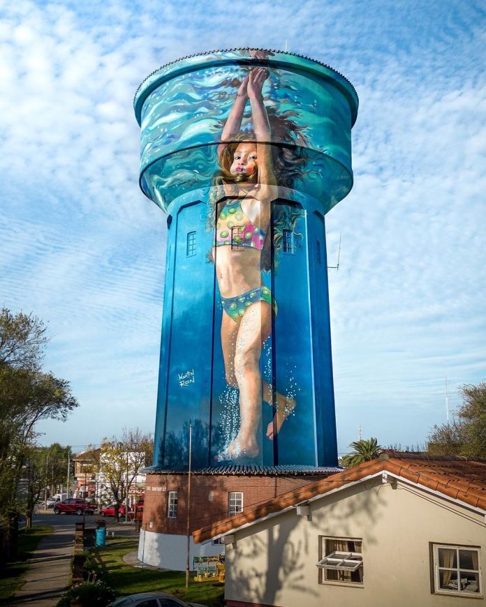 Another view of Martín Ron’s stunning mural in Miramar, Argentina, showcasing a young girl in a colorful polka-dot swimsuit diving headfirst into the water. Her hands are extended above her head, and her hair floats as if suspended in water, creating a dynamic and immersive underwater effect. The mural, painted on a 35-meter-high water tank, features vibrant blue hues and intricate detailing, making the entire structure appear as if it’s filled with water. The idyllic neighborhood and bright sky in the background amplify the captivating realism of the scene.
