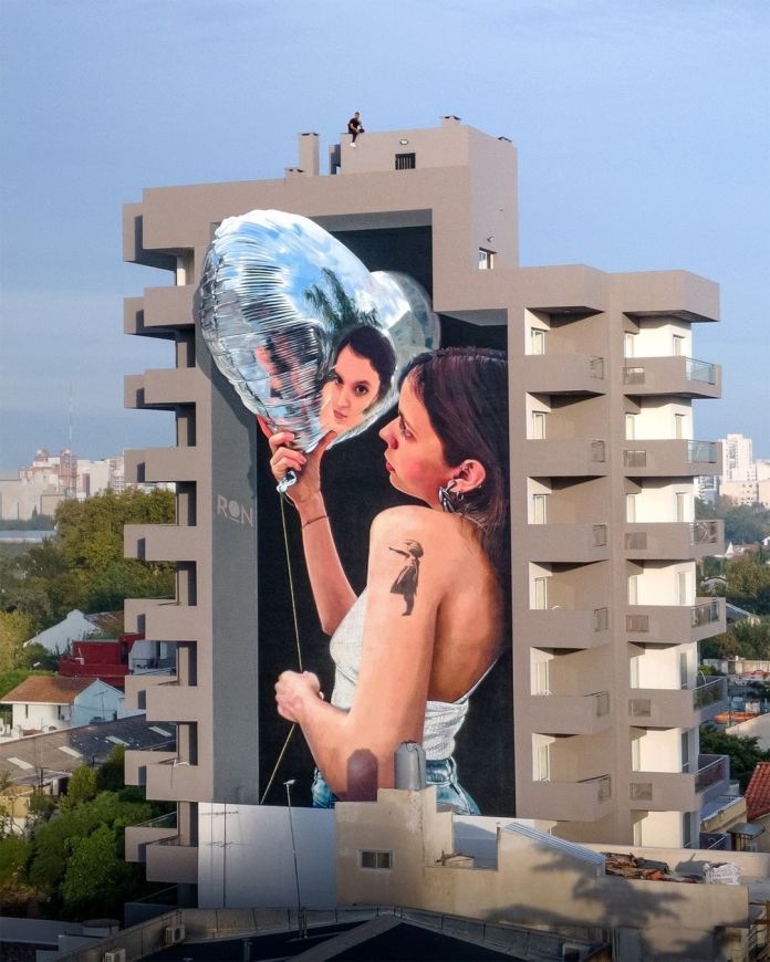 A captivating mural by Martín Ron in Bernal, Buenos Aires, Argentina, depicting a young woman holding a shiny, reflective heart-shaped balloon. The balloon mirrors her face, creating a mesmerizing effect that blends realism and surrealism. The woman, wearing a white tank top and denim jeans, has a small tattoo of a silhouette on her upper arm, adding a personal touch to the artwork. The mural is painted on the side of a modern high-rise building, with neutral tones that contrast beautifully with the vibrant details of the artwork. The urban surroundings and soft blue sky enhance the mural's visual impact.