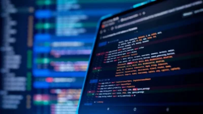 Close-up of a tablet displaying colorful code on a dark background, with blurred lines of programming visible behind it.