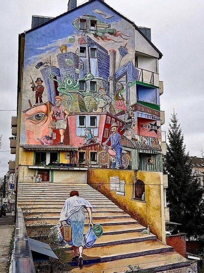 Large mural by Klaus Klinger in Düsseldorf, Germany, showing a surreal cityscape with giant faces, cyclists, and imaginative figures painted across a building, with staircases blending into the real street.