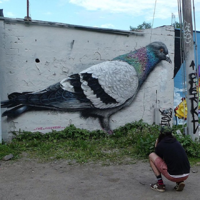 Mural by The Highness in Stockholm, Sweden, showing a giant pigeon painted with detailed feathers and iridescent colors against a white wall, with a person crouched below for scale.