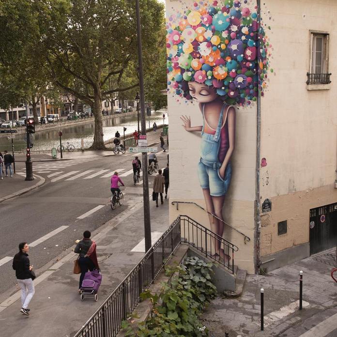 Mural in Paris by Vinie Graffiti showing a girl in blue overalls with a large afro made of colorful painted flowers, located on the corner of a building above a street staircase.