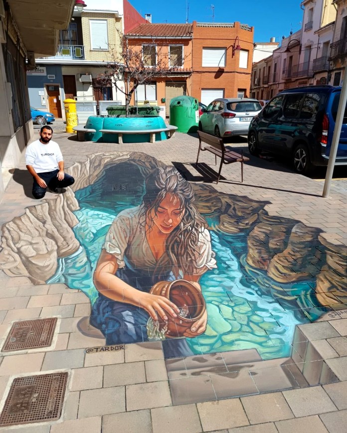 3D mural on a plaza in Riola, Spain showing a woman collecting water from a stream, painted on tiles by Juandres Vera and Tardor using realistic shadow and perspective techniques.