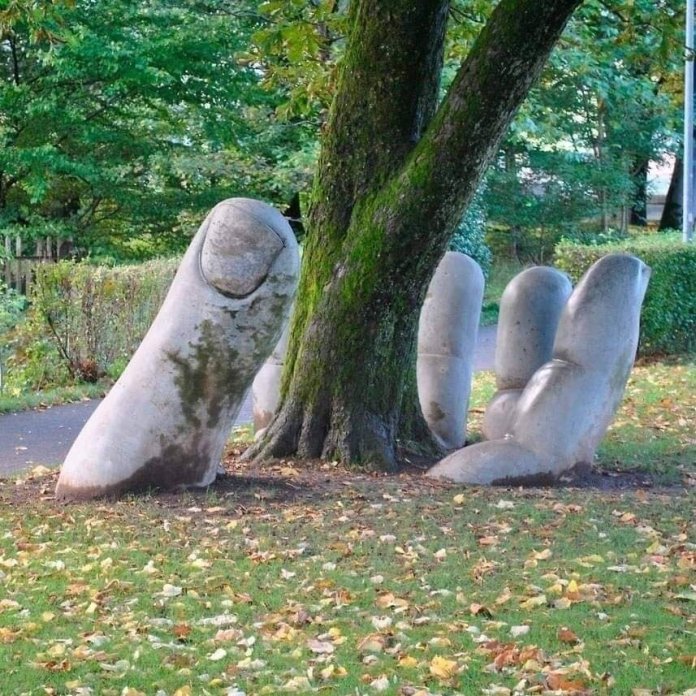 A large stone sculpture shaped like a hand rises from the ground in Glarus, Switzerland, gently cradling a tree trunk. The artwork symbolizes harmony between humans and nature, surrounded by lush greenery and fallen autumn leaves.
