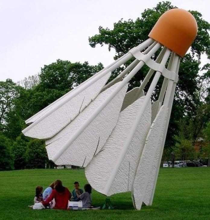 A massive white and orange shuttlecock sculpture lies on its side on a grassy field, creating the illusion of a playful, oversized badminton game in Kansas City, Missouri.