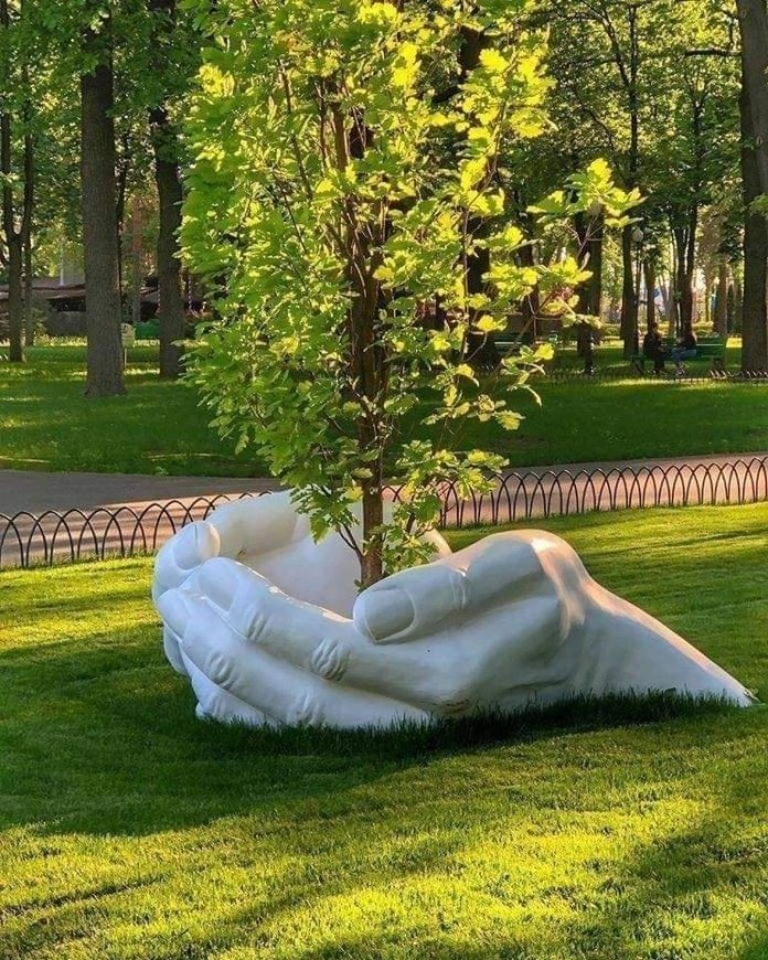 A white sculpture of two giant hands holding a young tree emerges from the grass in a serene park. Sunlight filters through the leaves, highlighting the peaceful connection between art and nature.