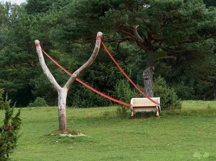 A creative outdoor sculpture shows a wooden bench attached to a giant slingshot made from tree branches and red straps, set in a green park surrounded by trees.