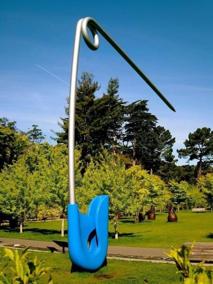 A giant blue and silver safety pin sculpture stands upright in a grassy park at the de Young Museum in San Francisco, surrounded by trees under a clear blue sky.