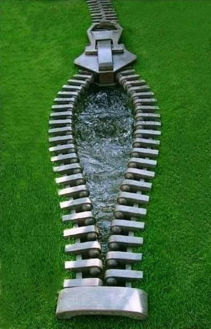 A unique sculpture shaped like a giant zipper appears to open the grassy earth, revealing a flowing stream of water beneath, blending creativity with natural elements.