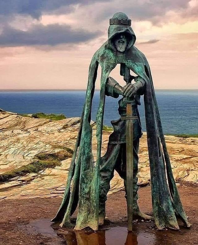 Statue of King Arthur by artist Rubin Eynon on the rugged cliffs of Tintagel, Cornwall, UK. The bronze figure stands cloaked and mysterious, with gaps in the metal giving a ghostly, ethereal appearance. Arthur holds the hilt of a sword, symbolizing his legendary status and connection to British folklore. The dramatic coastline and the vast expanse of the ocean serve as a backdrop, adding to the mythic atmosphere of the sculpture.
