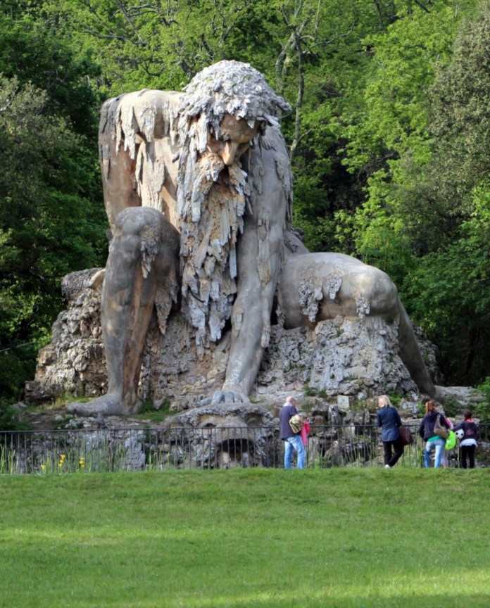 ‘Colosso dell'Appennino,’ an imposing 16th-century sculpture by Giambologna, located in the gardens of Villa Demidoff in Pratolino, Italy. The colossal figure, representing the Apennine mountain god, appears to emerge from the natural rock, his body weathered and adorned with moss and lichen, blending seamlessly with the surrounding greenery. Visitors walking nearby highlight the immense scale of the statue, which stands as a blend of art and nature, symbolizing the connection between humanity and the environment.