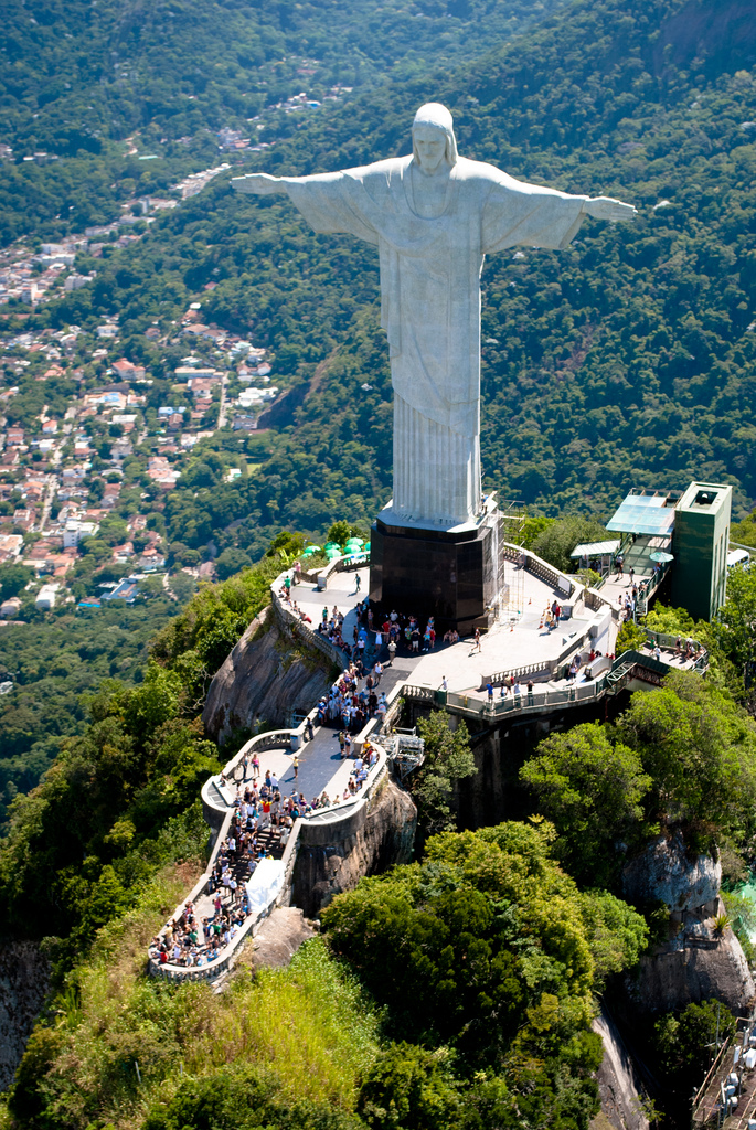 An aerial view of the iconic Christ the Redeemer statue in Rio de Janeiro, Brazil, overlooking the lush, mountainous landscape and the city below. The massive statue, with outstretched arms, stands atop the Corcovado Mountain, attracting numerous visitors seen gathering at the observation platform around its base. The vibrant green foliage contrasts with the grey stone of the statue, symbolizing peace and welcoming people from all over the world to admire the stunning views of Rio.