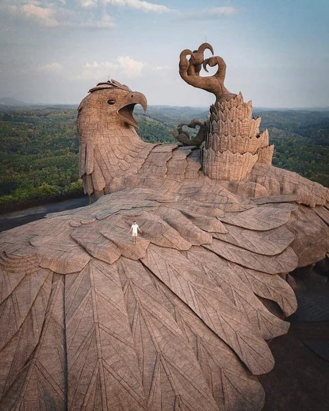A colossal stone sculpture of a bird with expansive wings spread across the landscape, known as the Jatayu Earth's Center in Kerala, India. This massive sculpture, representing the mythical eagle Jatayu from the Hindu epic Ramayana, is intricately detailed with feather patterns across its wings. The eagle's head is raised, its beak slightly open, and one talon is raised gripping a symbolic figure. A tiny human figure stands on one of the wings, emphasizing the immense scale of this artwork. Lush green forests surround the structure, blending nature with mythology.