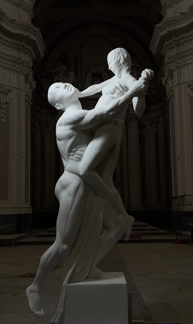 marble sculpture, Ajax and Cassandra, was crafted in 2022 by renowned Italian contemporary artist Jago. A powerful marble sculpture of two figures in an intense, intertwined embrace. The male figure, with a muscular form, holds the female figure, who appears to be floating or lifted. The female figure reaches up, her hand clasping the man’s shoulder as they gaze into each other’s eyes. The sculpture captures a moment of passion and strength, with intricate details highlighting the tension and connection between the figures. Set in an ornate interior with grand architectural elements, the artwork stands as a striking representation of human emotion and physicality.