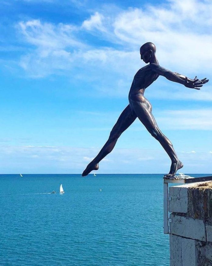 The Great Challenge by Nicolas Lavarenne in Antibes, French Riviera. This striking bronze sculpture depicts a dynamic, elongated figure balancing on the edge of a cliff, arms outstretched as if preparing to leap into the vast blue sea below. The figure’s athletic stance and poised energy contrast against the serene backdrop of the ocean and sky, symbolizing courage and the thrill of taking risks. Sailboats dot the horizon, adding a sense of scale and tranquility to the dramatic scene.
