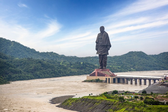 The Statue of Unity in India, the tallest statue in the world at 182 meters (597 feet) tall, towering over the surrounding landscape with lush green hills and a river nearby. This monumental statue of Sardar Vallabhbhai Patel, a key leader in India’s independence, stands on a massive platform with an observation deck, emphasizing its grandeur and scale. The statue, nearly three times the height of the Statue of Liberty, represents unity and strength, with a bridge connecting it to the nearby complex and viewing areas.