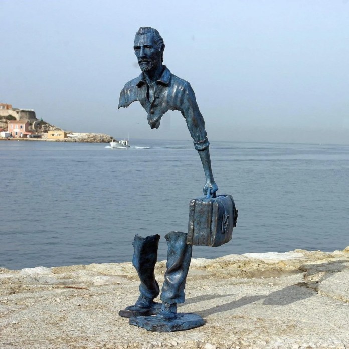 Sculpture by Bruno Catalano depicting a partially absent human figure holding a suitcase. The sculpture, positioned near a waterfront, portrays a man with significant portions of his body missing, creating a surreal effect as the head, upper torso, and legs appear disjointed yet balanced. The artwork symbolizes themes of travel, identity, and the idea of leaving parts of oneself behind. The backdrop includes a serene view of the sea, with distant buildings and a boat in the background.