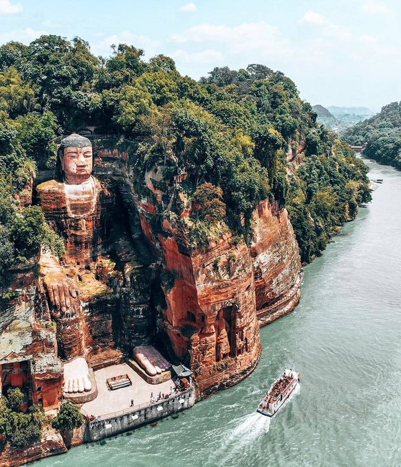 The Leshan Giant Buddha, an enormous 71-meter (233 ft) tall stone statue carved into the cliffside in Leshan, Sichuan, China, overlooking the convergence of three rivers. Created in 800 AD, this ancient Buddha statue is the tallest pre-modern statue in the world. Surrounded by lush greenery and set against red sandstone cliffs, the Buddha sits serenely with hands resting on his knees, while a boat passes below, illustrating the scale of this monumental figure. The scene reflects the blend of natural and cultural heritage in this iconic location.