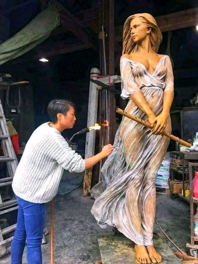 Artist Luo Li Rong working on a life-sized, hyper-realistic sculpture of a woman. The sculpture is meticulously crafted, capturing fine details of the figure’s delicate features and flowing draped garment, which clings to her form as if made of sheer fabric. Luo uses a torch to carefully refine parts of the sculpture, demonstrating her precise technique. The studio setting and surrounding tools emphasize the artist’s skill and dedication to her craft. Luo Li Rong is known for her highly realistic clay and bronze sculptures that celebrate the grace and beauty of the human figure.