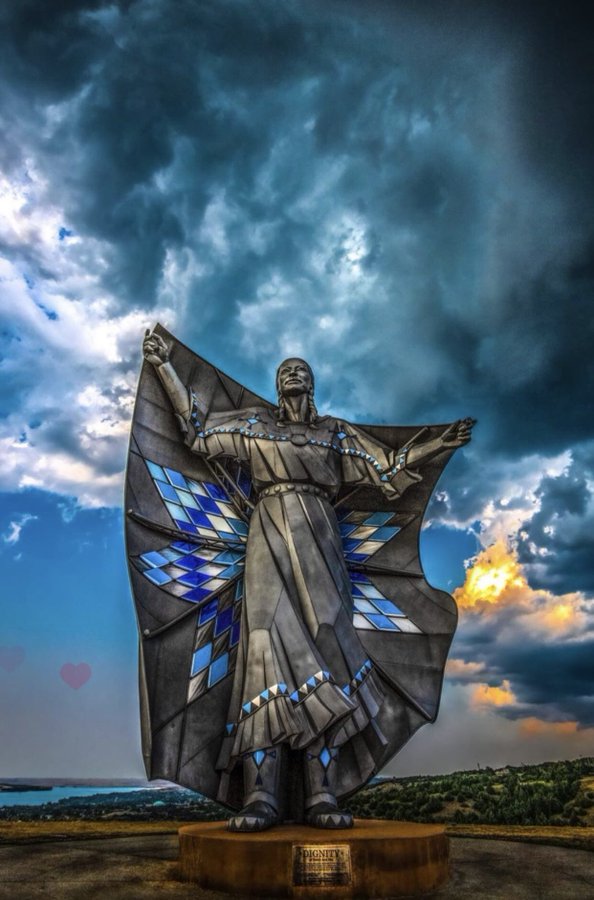 The Dignity statue in South Dakota, a stunning 50-foot-tall sculpture of a Native American woman draped in a star quilt. The statue features a blend of metal and vibrant blue diamond shapes that catch the light, symbolizing the cultural heritage and spirit of the Lakota and Dakota people. The woman’s expression is serene, with her arms outstretched as if embracing the landscape around her. Dark clouds and sunlight create a dramatic sky backdrop, enhancing the sculpture’s powerful presence.