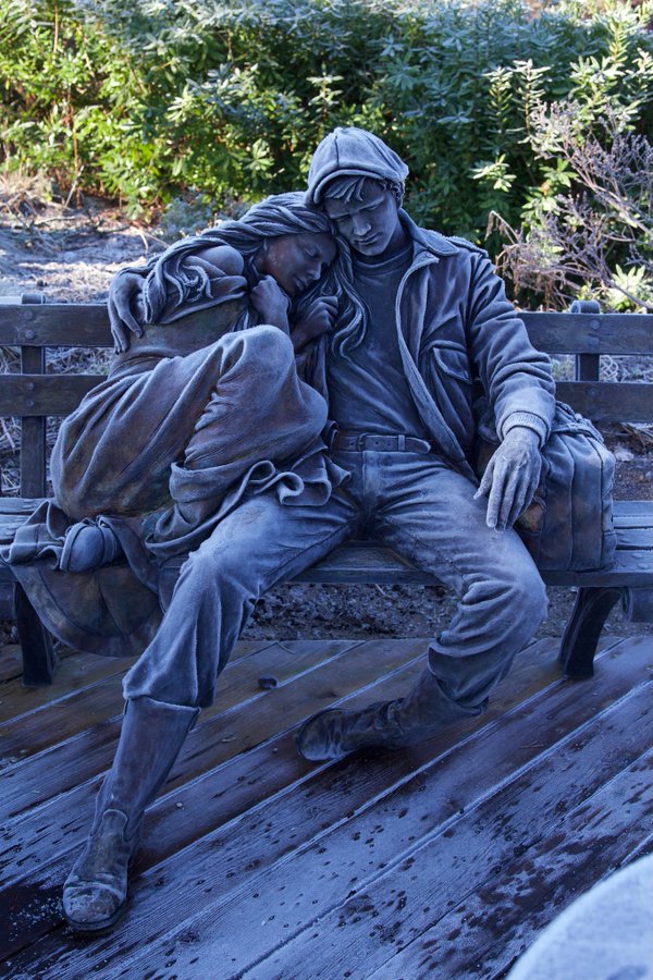 "Departure," a captivating bronze sculpture by artist George Lundeen, depicting a young man and woman resting on a bench together. The man wears a jacket and cap, while the woman leans on his shoulder with her eyes closed, wrapped in a blanket for warmth. The sculpture is subtly highlighted by a layer of frost, enhancing the scene with a sense of cold and quiet intimacy, as if capturing a moment of departure or waiting. The frost-covered bench and wooden floor add texture and depth to the atmosphere around the figures.