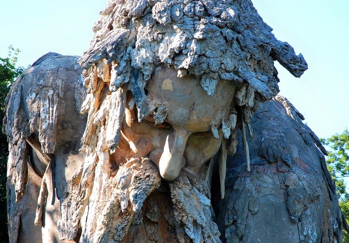 Close-up view of the Apennine Colossus, a 16th-century sculpture by Giambologna in the Villa Demidoff park near Florence, Italy. This detailed shot highlights the weathered face of the colossal figure, with a prominent nose and cascading beard that merges into the surrounding rocky textures. The intricately sculpted hair, resembling rough stone and vegetation, adds to the illusion of the sculpture blending seamlessly with its natural environment. Sunlight illuminates the aged surface, showcasing the fine craftsmanship and enduring beauty of this Renaissance masterpiece.