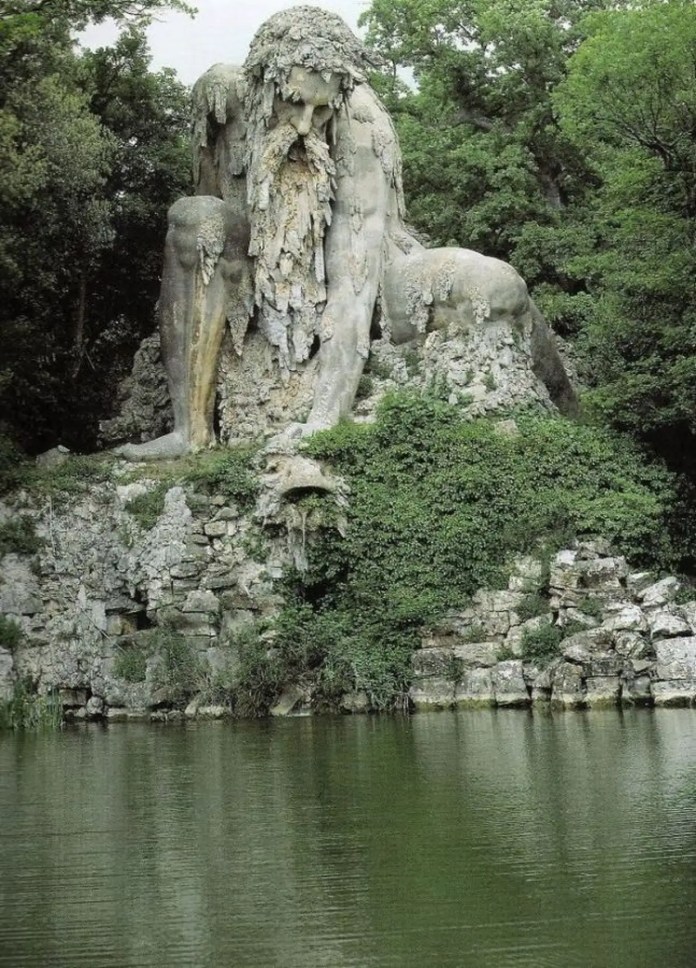 Full view of the Apennine Colossus, a monumental 16th-century sculpture by Giambologna located in the Villa Demidoff park near Florence, Italy. The massive stone figure represents a crouching mountain god or titan, with a rugged, weathered surface that mimics the textures of rock and vegetation. The sculpture is partially covered by creeping greenery, blending seamlessly into the surrounding natural landscape. A calm pond in the foreground reflects the lower portion of the sculpture, enhancing the serene and majestic atmosphere of this Renaissance masterpiece.
