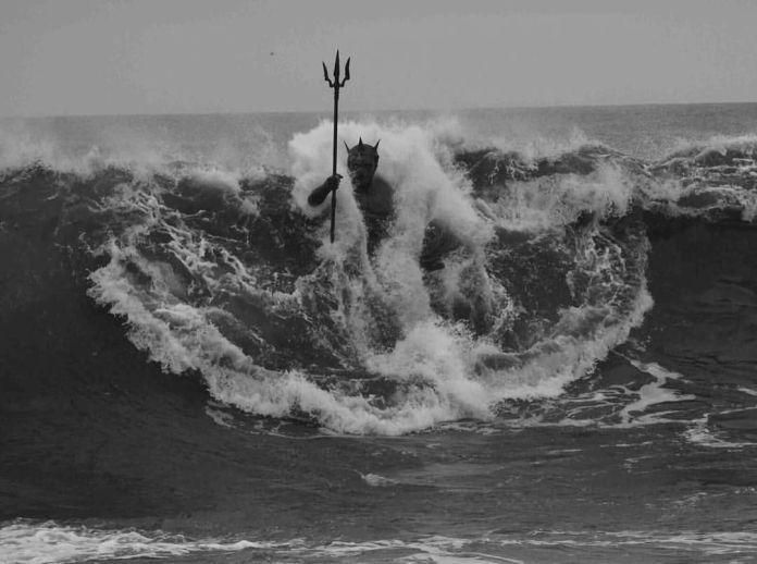 A black-and-white photograph of Neptune, or Poseidon, a powerful ocean sculpture in Gran Canaria, Spain. The statue appears to rise dramatically from a massive wave, holding a trident aloft, as if commanding the sea. The monochrome effect highlights the intensity of the waves and the mythological figure's presence amidst the churning ocean.