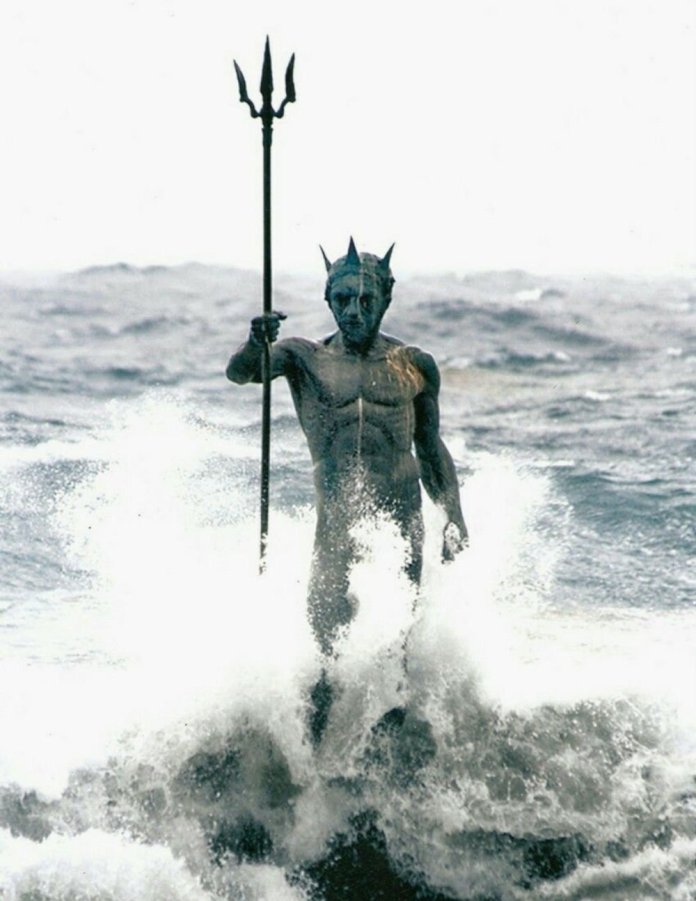 A dramatic ocean sculpture of Neptune, also known as Poseidon, standing powerfully amidst crashing waves in Gran Canaria, Spain. The statue, featuring a muscular figure with a trident and crown, emerges from the sea as waves break against its base, creating a striking and dynamic scene of mythology and nature colliding.